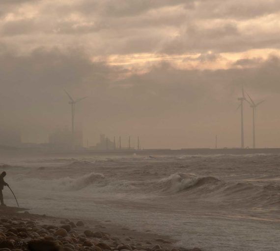A man at the shore stands in a storm with wind turbines behind him