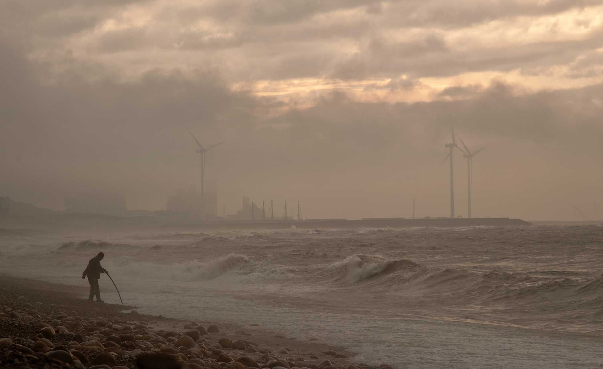A man at the shore stands in a storm with wind turbines behind him