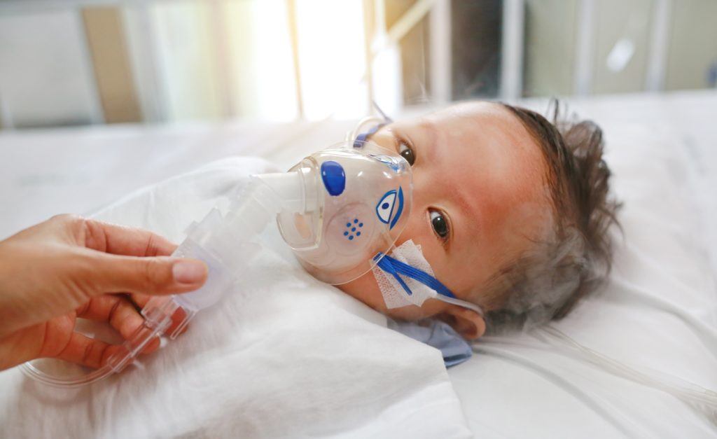 Sick baby boy applying inhale medication by inhalation mask to cure Respiratory Syncytial Virus (RSV) on patient bed at hospital.