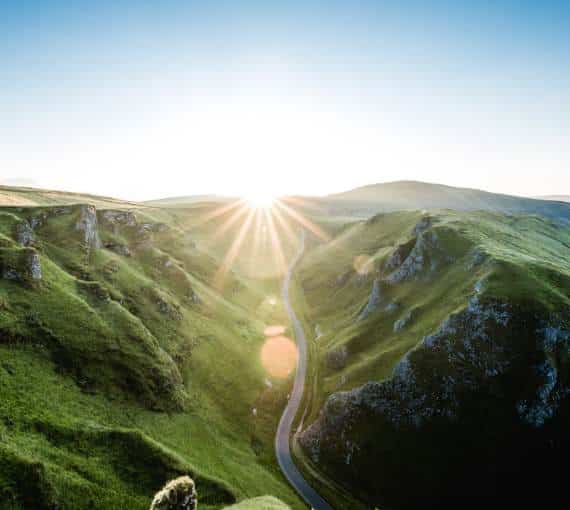 The sunrise and sun beams over green mountains and a winding road.