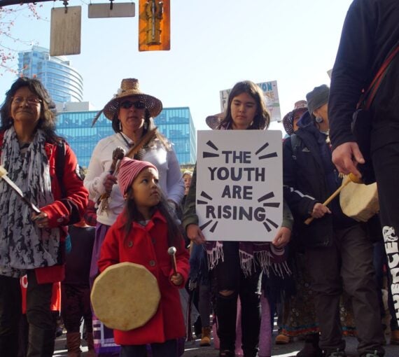 Youth and Indigenous People at a rally holding a sign that reads 'the youth are rising'